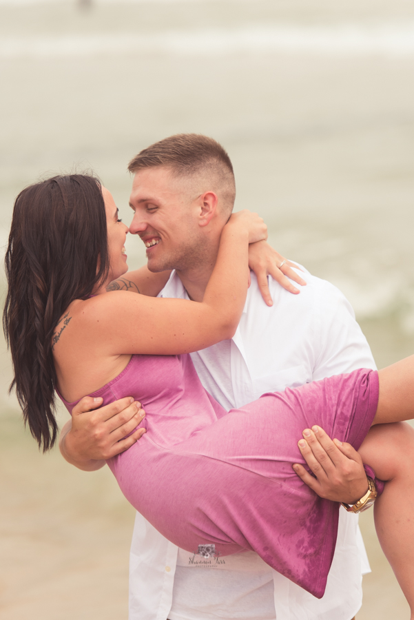 Engagement Photography in Ludington MI of guy standing on beach holding his bride to be