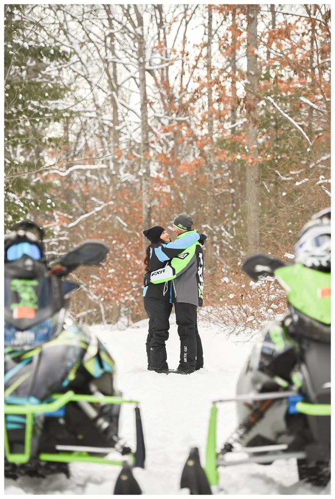 couple standing in snow holding each other behind snowmobiles
